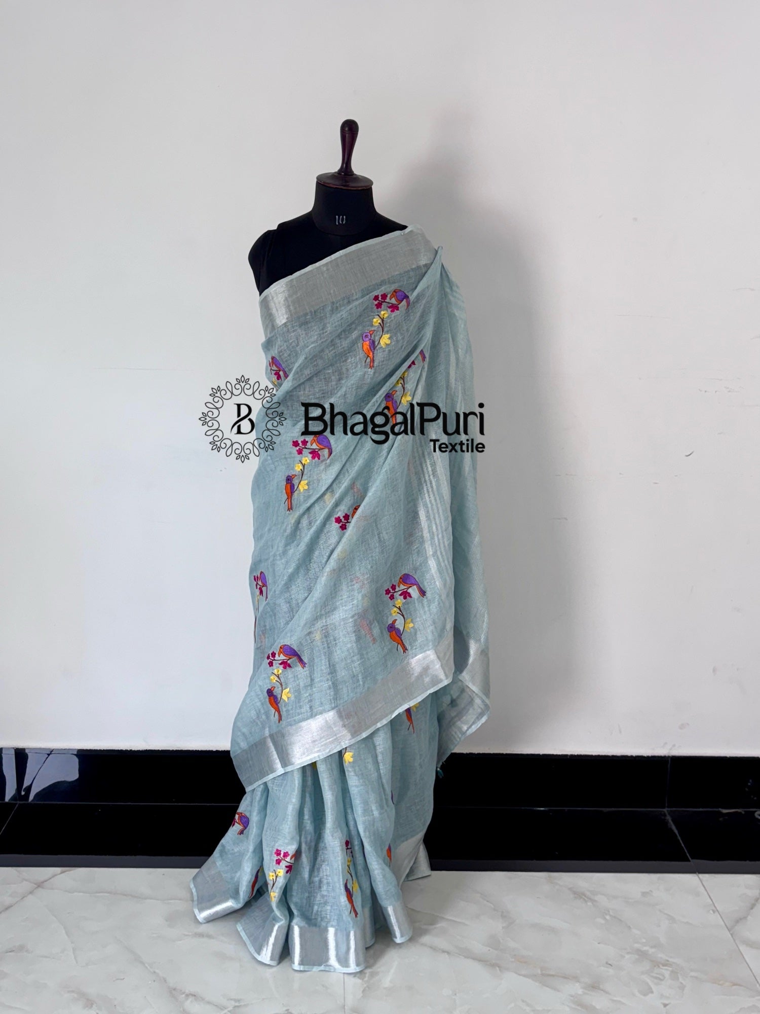 Light blue saree with floral patterns on a mannequin against a white background, branded 'Bhagalpuri Textiles'.
